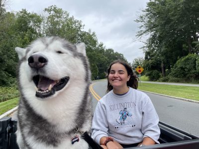 Picture of Erin McKeehan and Jonathan XIV, the husky canine mascot for UConn. Erin is sitting in the back of a gator utility car with the dog and smiling while the dog has his eyes closed happily with the wind in his fur.