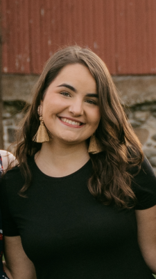 Picture of Erin McKeehan from the waist up. Erin is smiling while wearing a solid black short sleeve sundress and tan tassel earrings. In the background, you can see part of a wall of a red barn sitting on top of a stone foundation. 